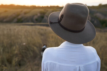 Back view of faceless woman in big hat enjoying warm sunset at hillside meadow
