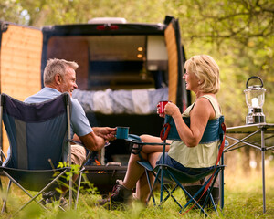 Rear View Of Senior Couple Camping In Countryside With RV Drinking Coffee By Outdoor Fire
