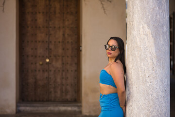 Fototapeta premium Young, beautiful, brunette, South American woman in blue dress and sunglasses posing calm and relaxed leaning on a marble column. Concept beauty, fashion, trend, peace, tranquility.