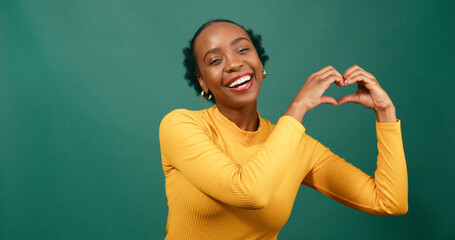 Young Black woman forms heart with hands to side, smiling green studio