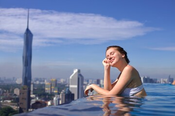 Fototapeta premium Portrait of happy young woman in swimming pool on the rooftop of skyscraper hotel with beautiful city urban view
