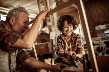 Senior male carpenter teaching his grandson how to use a hammer in a carpentry shop - obrazy, fototapety, plakaty