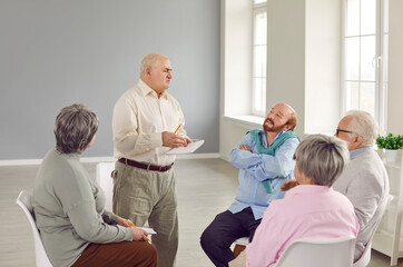 Obraz premium Mature man talks about his problems and experiences to group of other seniors during group therapy for elderly. Group of senior people sitting in circle during therapy session at retirement home.