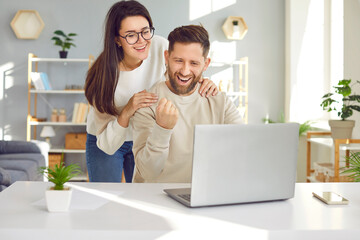 Cheerful young couple using laptop computer at home. Excited young couple feeling happy getting amazing sale offer, good email, surfing internet or purchasing online