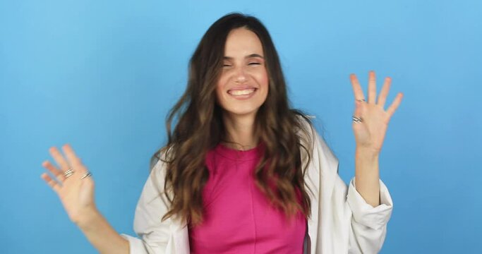 Woman in pink top, white shirt dancing, showing victory, v sign. Funny unusual brunette woman hair having fun, hands up, grimaces, smiling, dancing in studio against blue background. Music, dance.