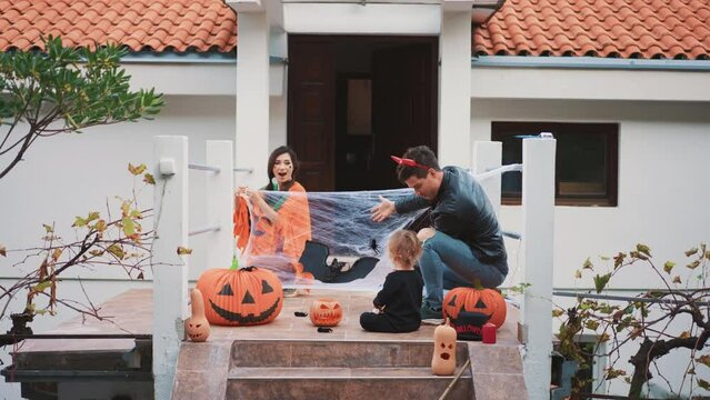 Joyous happy mix race Asian family decorate porch smiling and laughing while hanging bets and spiders, carved pumpkins on Halloween. Dad mother cute baby celebrate autumn tradition holiday together