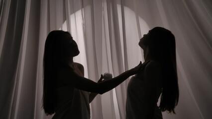 Medium side view shot of two sisters, young women's silhouettes wrapped in a towel. One of them is applying body cream on the other one's skin in a muffled light.