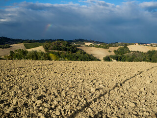 Scenic view of tilled soil with rolling hills, trees and a partial rainbow in a dark sky on a sunny day in San Procolo, Monte Vidon Combatte in the Marche Region; Province of Fermo, Italy