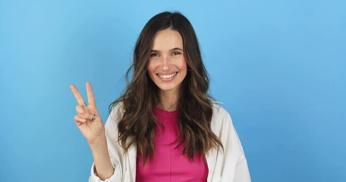 Young positive woman showing peace gesture, expressing fun and happiness, sign of victory, blue studio background. Young caucasian woman showing victory sign and smiling broadly.