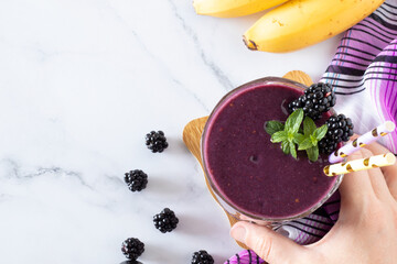 Hand-holding blackberry banana smoothie with fresh mint in a glass with eco-friendly straws on...