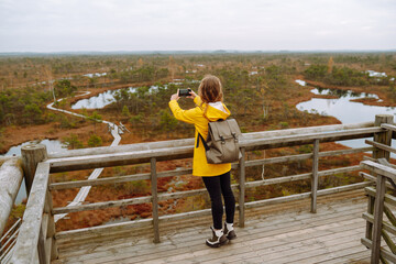 Smiling traveler in a bright coat with a backpack walks along a wooden path in the swamps. Travel...
