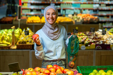 Portrait of a female buyer in a supermarket, a Muslim woman in a hijab is smiling happy and looking at the camera, choosing apples and fruits in a large grocery store.