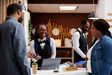 Smiling positive African American woman front desk agent receiving contactless payment from guests at reception counter. Black couple of tourists paying for room with credit card at check-in