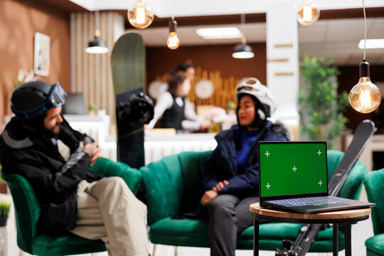 Youthful couple with snow gear in hotel lobby engage in conversation as laptop having green screen display positioned on table. Image shows device with mockup template and guests seated in ski resort.