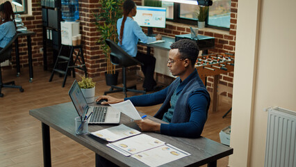 Corporate worker reviewing paperwork in office, looking at documents with statistics files in coworking space. African american man working on business development with data analytics.