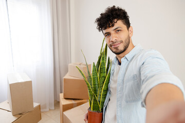 young Hispanic man taking a selfie in his new house surrounded by movers - young man taking photos...