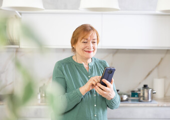 Mature woman sitting in kitchen is talking via video link using messenger.Lady is holding phone in hands and writing post on social network