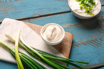Board with bowls of tasty sour cream and green onion on blue wooden background