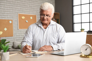 Senior man with laptop writing in notebook at home