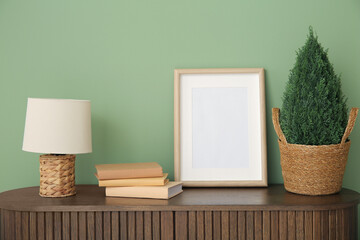 Blank frame, books, lamp and tree in pot on wooden cabinet near green wall