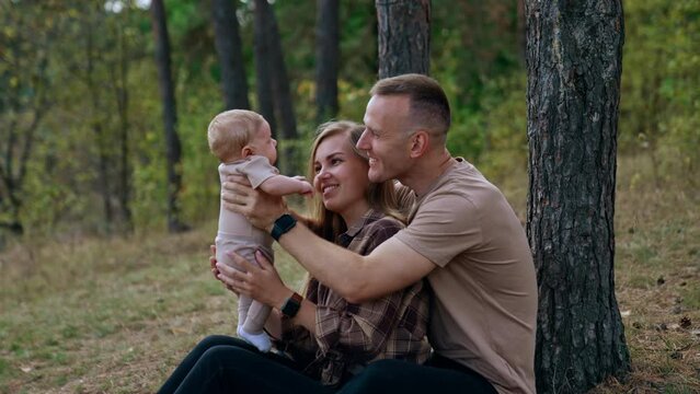 Caucasian parents sit in the woods holding their newborn child. Happy couple talk, smile and kiss their lovely infant.