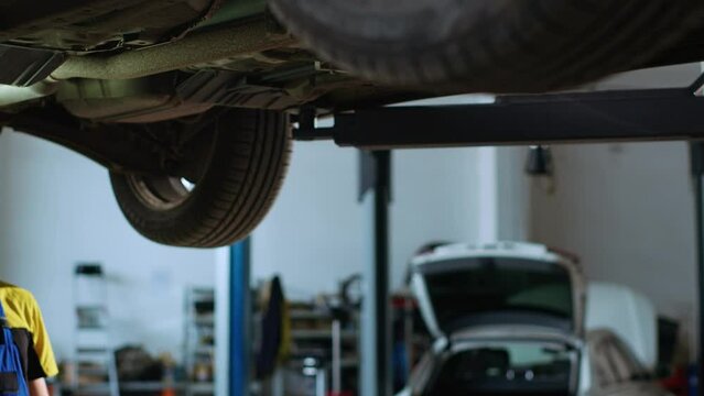 Precise mechanic underneath vehicle on overhead lift in garage, checking components. Employee using work light to make sure automotive underbody is in perfect condition, doing close examination