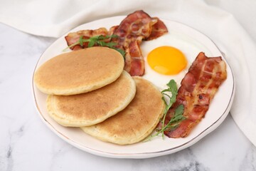 Plate with tasty pancakes, fried egg, arugula and bacon on white marble table, closeup