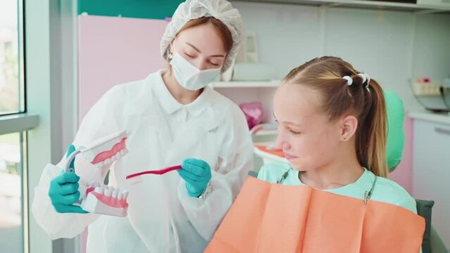 teengirl learning to brush teeth in modern dental clinic, doctor woman showing jaw model for child