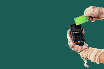 Mummy hands with credit card and payment terminal on green background. Halloween celebration