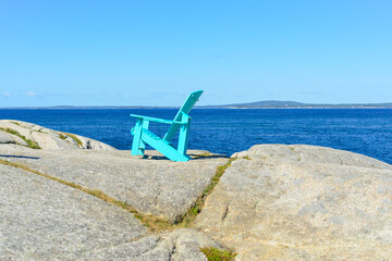 Fototapeta na wymiar A bright teal green and empty but sturdy resin material Adirondack chair on the rocky ground overlooking the ocean and an island. The terrain is textured molten pebbly rock. The sky is clear blue. 