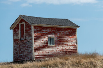 A small red wooden barn or shed in the middle of a grass covered field. There's a small hill on which the building sits. There's a hay loft door and a small glass window in the old historic building. 