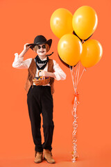 Little boy dressed for Halloween as cowboy with candies and balloons on orange background