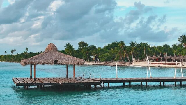 Wooden pier over turquoise sea waves against the backdrop of a white resort beach. Nature landscape of the ocean coast with palm trees. Caribbean sea and sky. Travel to a tropical paradise.