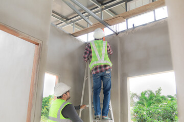Asian or Indian contractor man business partnership inspect ceiling room climbing ladder using measurement equipment with young foreman worker wearing safety white helmet, healthy men at work concept © Rakchanok