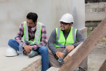 Two Asian or Indian builder workers wearing safety reflected vests and helmets tired thirsty hydrated drinking water from plastic bottles together relaxed sitting on construction site talking together