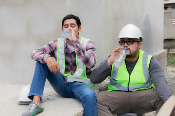 Two Asian builder workers wearing safety reflected vest and helmet tired thirsty hydrated drinking...
