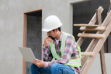 Asian or Indian foreman with beard wearing safety reflected vest sit on ladder outdoor at work construction site, thoughtful architect engineering middle-aged male working looking laptop technology