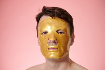 Face of mature man wearing golden hydrogel sheet mask, isolated on pink
