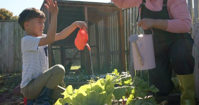 Senior biracial grandmother and grandson watering plants in sunny garden, slow motion