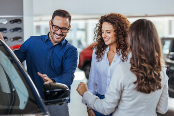 Couple looking at cars at the dealership and talking to the salesman - business concepts. Couple speaking about buying new car with auto salesman at vehicle dealership. 