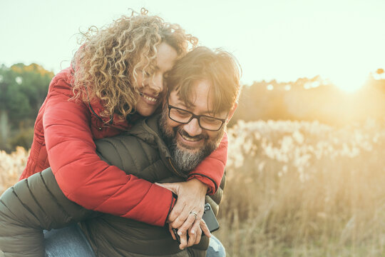 Young Adult Mature Couple In Love Hugging And Smiling With Romance Expression On Face. One Man Carry Woman In Piggyback On His Back. Female Bonding Male In Outdoor Leisure Activity Together. Happiness