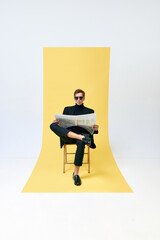 Handsome stylish man in black suit and sunglasses, writer, journalist reading newspaper against yellow studio background. Concept of human emotions, lifestyle, mass media, journalism, profession. Ad