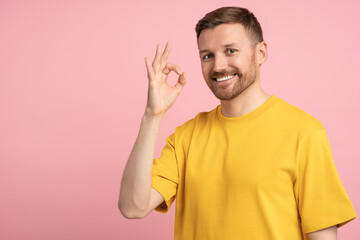 Positive happy man showing OK sign hand gesture on pink background with copy space looking at...