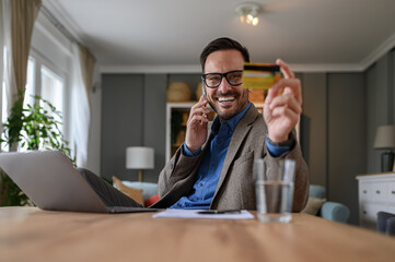 Handsome entrepreneur in elegant suit holding credit card and talking over smart phone at desk