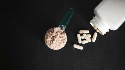Chocolate whey protein powder in measuring spoon, white capsules of amino acids, vitamins and...