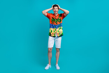 Fototapeta na wymiar Full size photo of impressed guy wear tropical shirt stylish shorts arms on head astonished staring isolated on blue color background