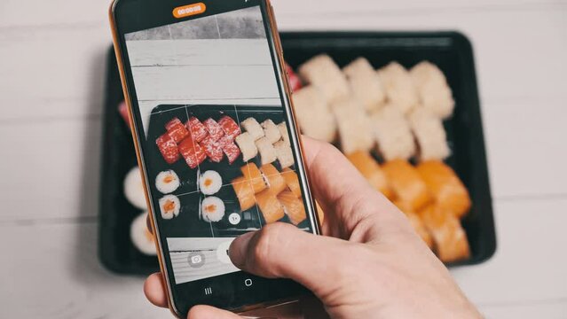 Blogger taking a video of sushi rolls by a smartphone camera for posting on social networks. Male hands filming Japanese food in a delivery plastic box on a mobile phone. Staged shot for social media.