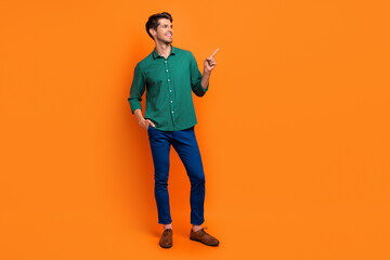 Full length photo of cute cheerful man dressed green shirt showing finger empty space isolated orange color background