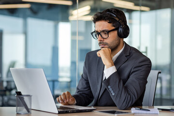 Fototapeta premium Serious thinking and concentrated man working with laptop inside office, man in headphones listening to audio books and podcasts at workplace.