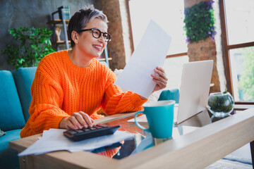 Photo of charming positive elderly lady wear orange pullover counting records modern device indoors apartment room